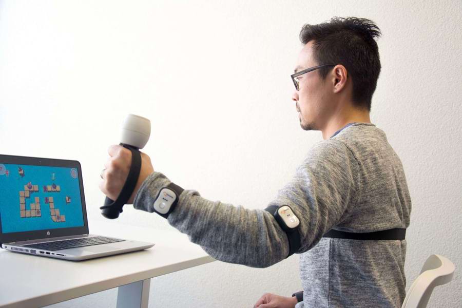 Mand sidder med sensorer på arme og bryst og holder en bevægelsessensor, mens han kigger på et spil på en skærm