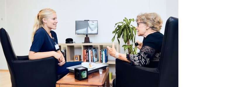 En ung kvinde sidder ved et sofabord og taler med en ældre kvinde. På bordet ligger MemoryCube-terningen