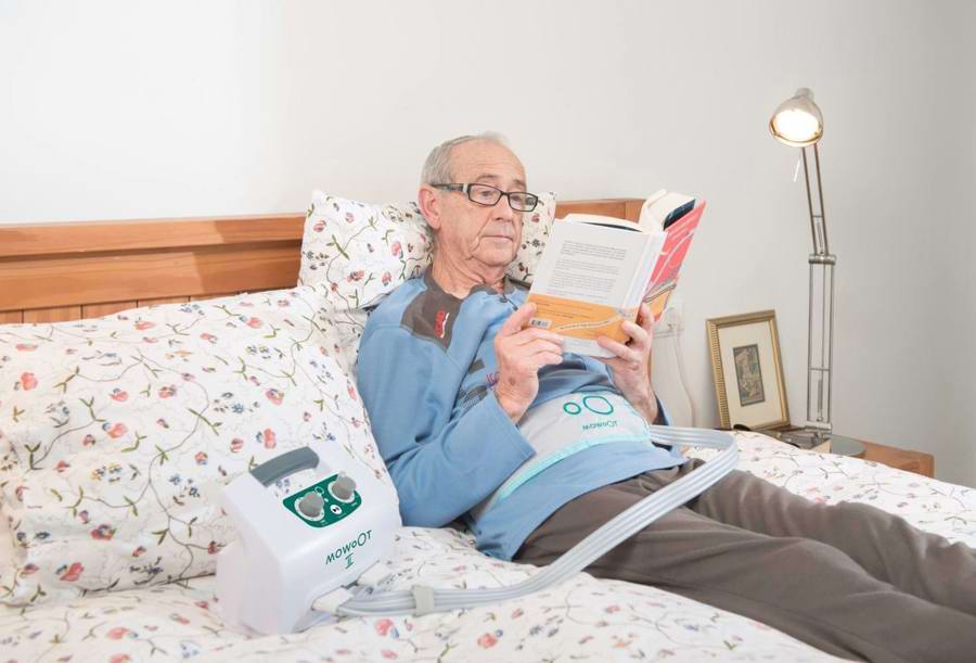 Ældre mand sidder med Mowoot bæltet på og læser i sin seng