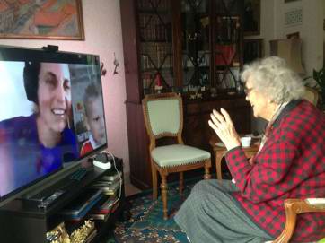 Ældre kvinde sidder i sin stue og taler med sin familie - på fjernsynsskærmen ses et billede af en ung kvinde og et barn