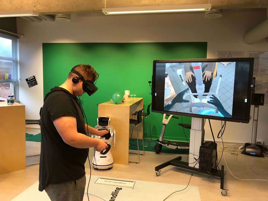 Ung mand med VR-briller og controllere. På skærmen bag ham kan man se en rollator i en virtuel verden
